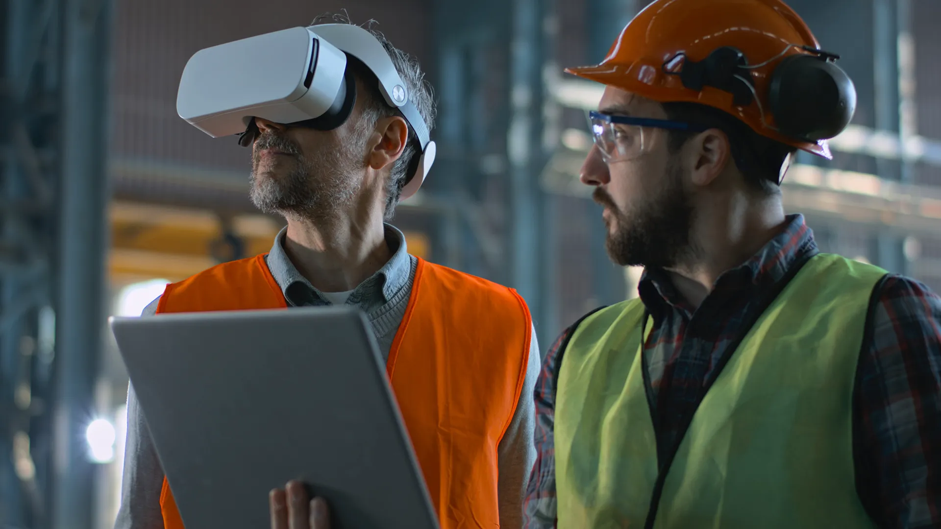 Two professional industry technicians wearing safety uniform and hardhats stand at manufacturing factory, use laptop and AR headset. Mature engineer and adult employee work in warehouse.